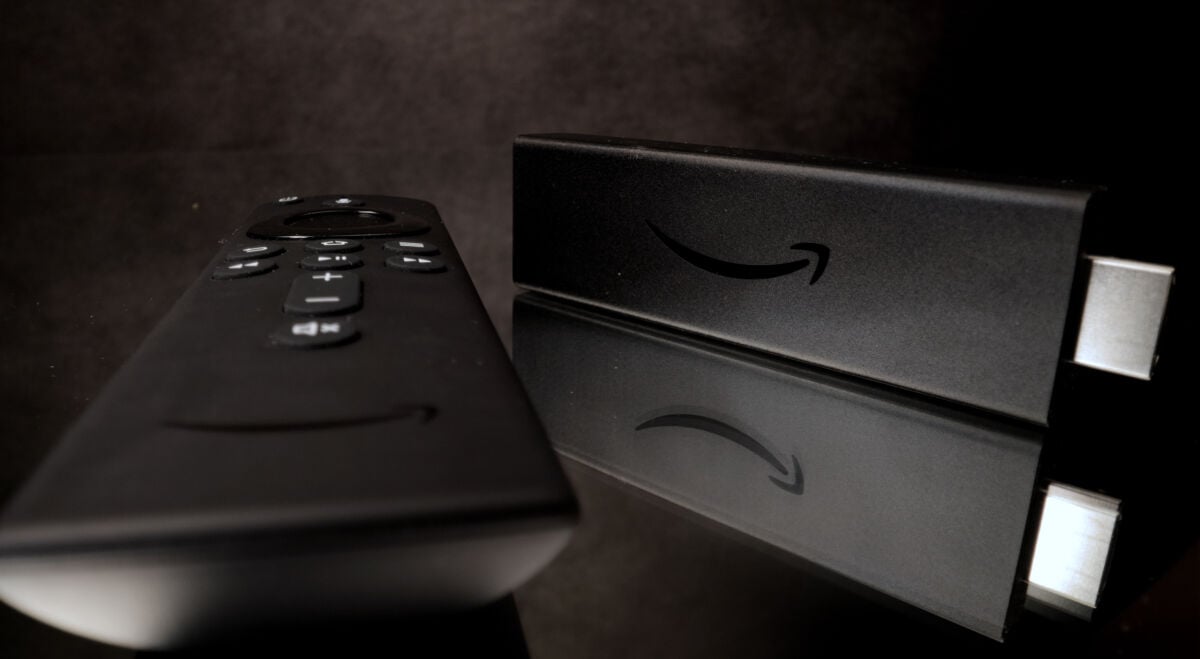 Amazon의 Fire TV Stick 리모컨과 동글이 어두운 배경에 배치되어 있으며 Amazon의 스마일리 로고가 장치와 리모컨에 표시되어 있습니다.